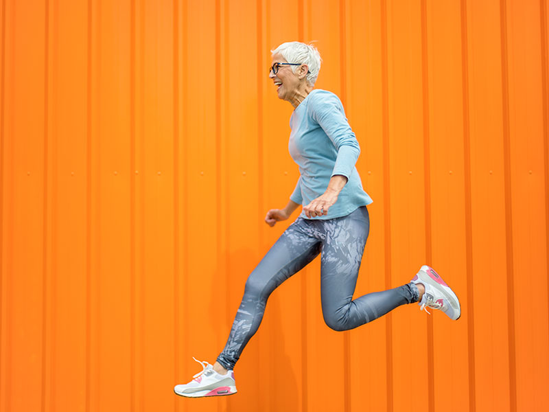 Woman in leggings and athletic footwear jumping and laughing against a bright orange background.