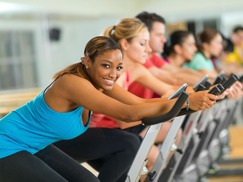 Women exercising on exercise bikes. The woman in front is smiling at the camera. Her energy levels are at a peak due to supplementing her diet with CoQ10.
