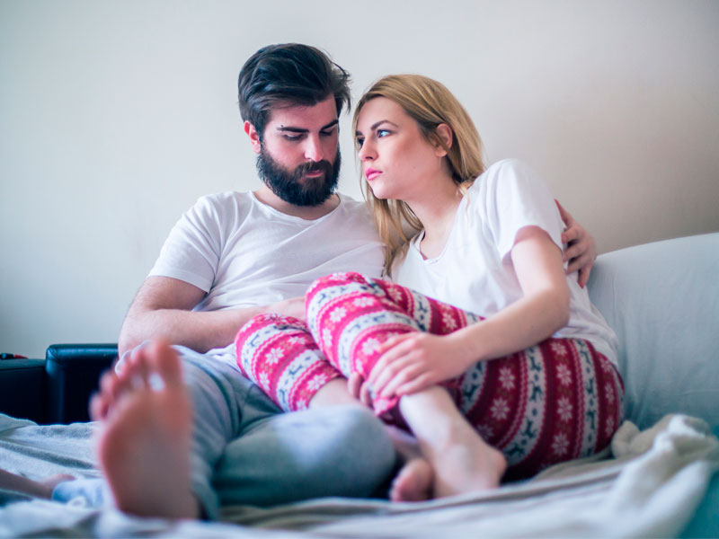 Couple sitting on bed looking sad and discouraged due to problems with infertility. The man is comforting the woman.